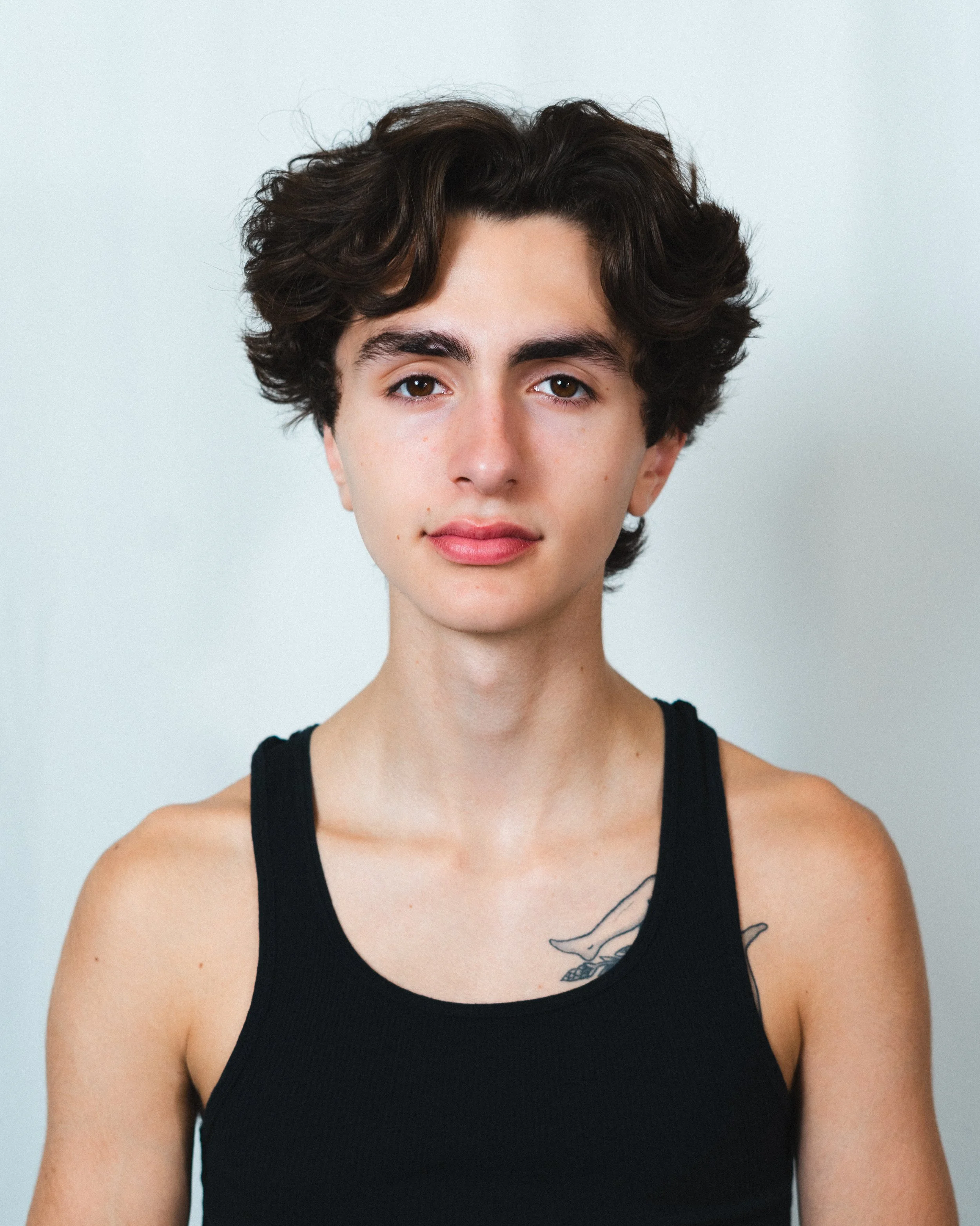 portrait of a young man with short curly brown hair wearing a black tank top, standing against a plain white background
