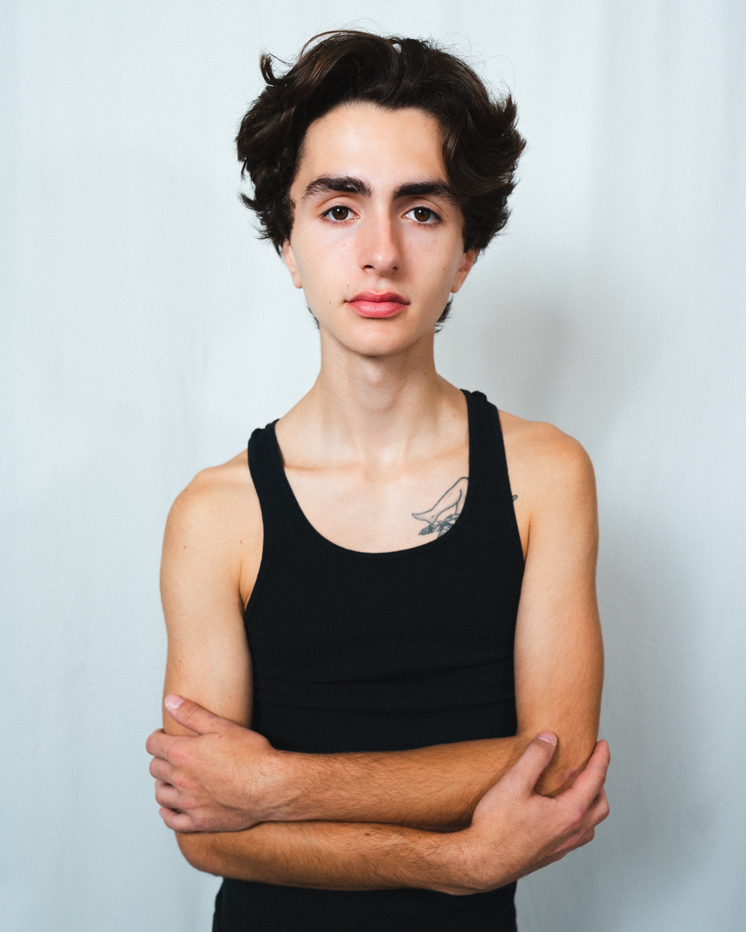 Portrait of a young person with short dark hair, wearing a black tank top, crossing arms, and standing against a plain light background.