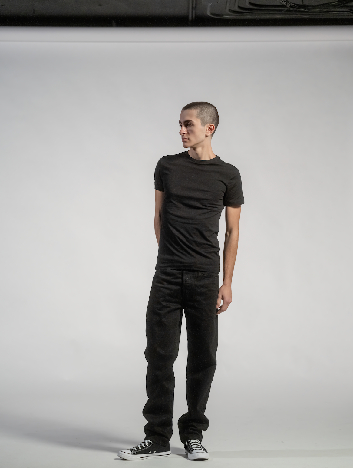 A young man with short hair wearing a black t-shirt, black pants, and black Converse sneakers standing in front of a white background.