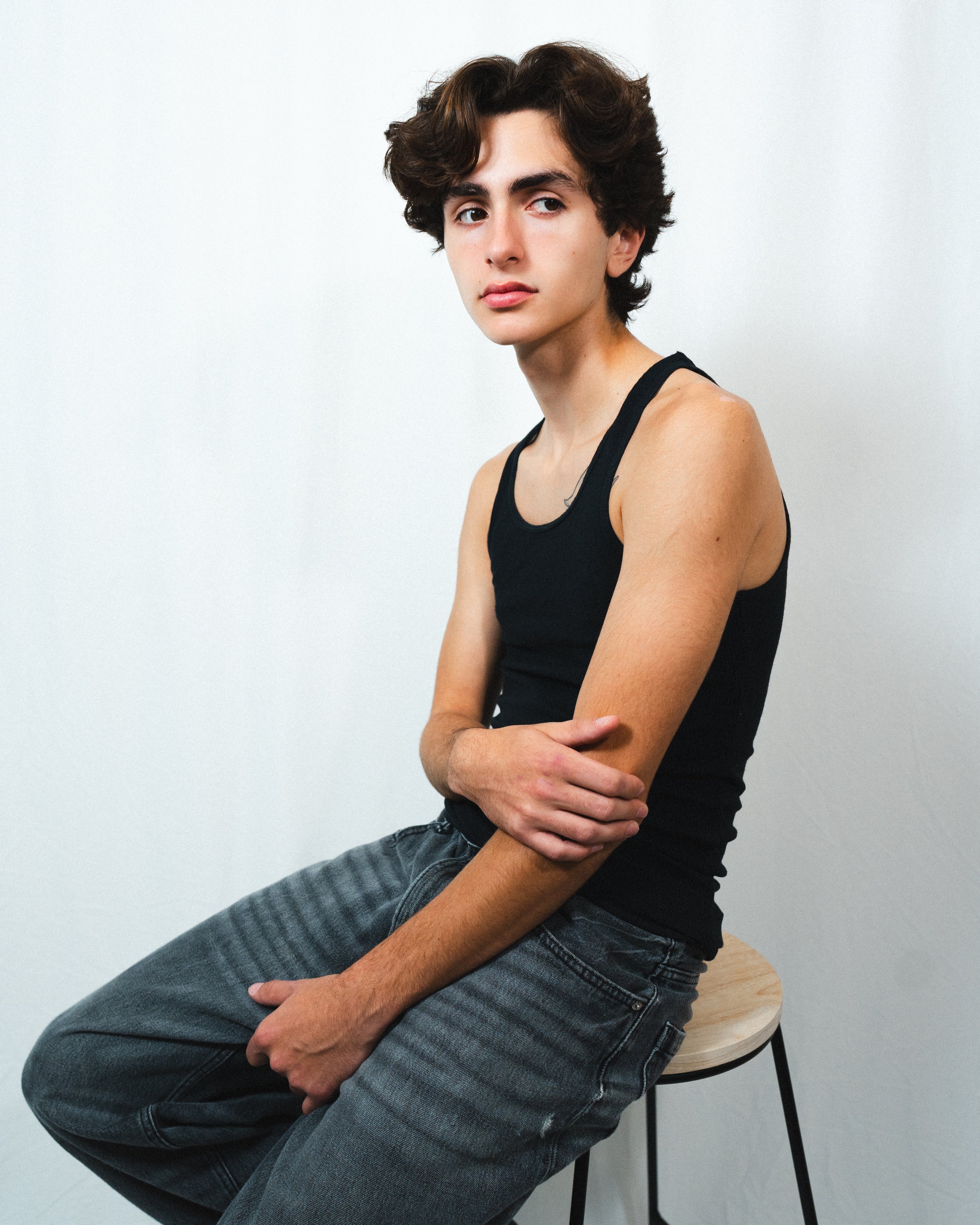 A young man with curly brown hair sitting on a wooden stool, wearing a black tank top and gray jeans, against a plain white wall.