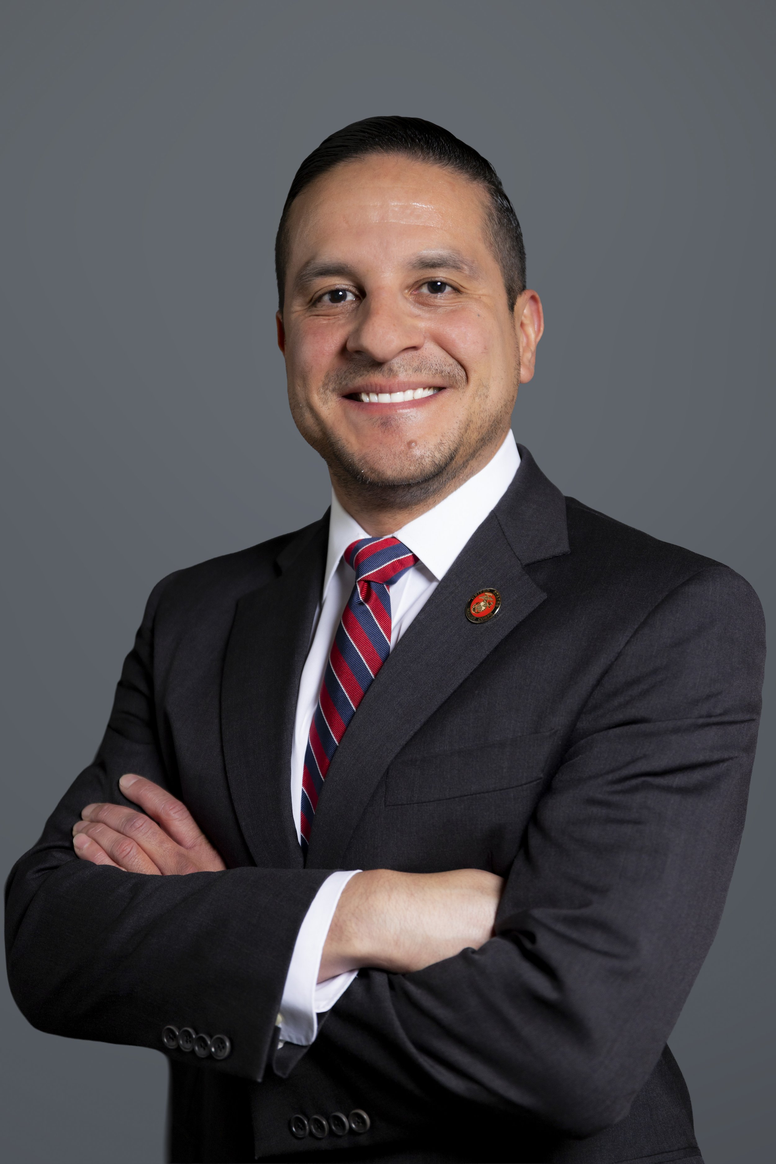 A man in a dark suit, white shirt, and striped red and blue tie, smiling with arms crossed, against a gray background.