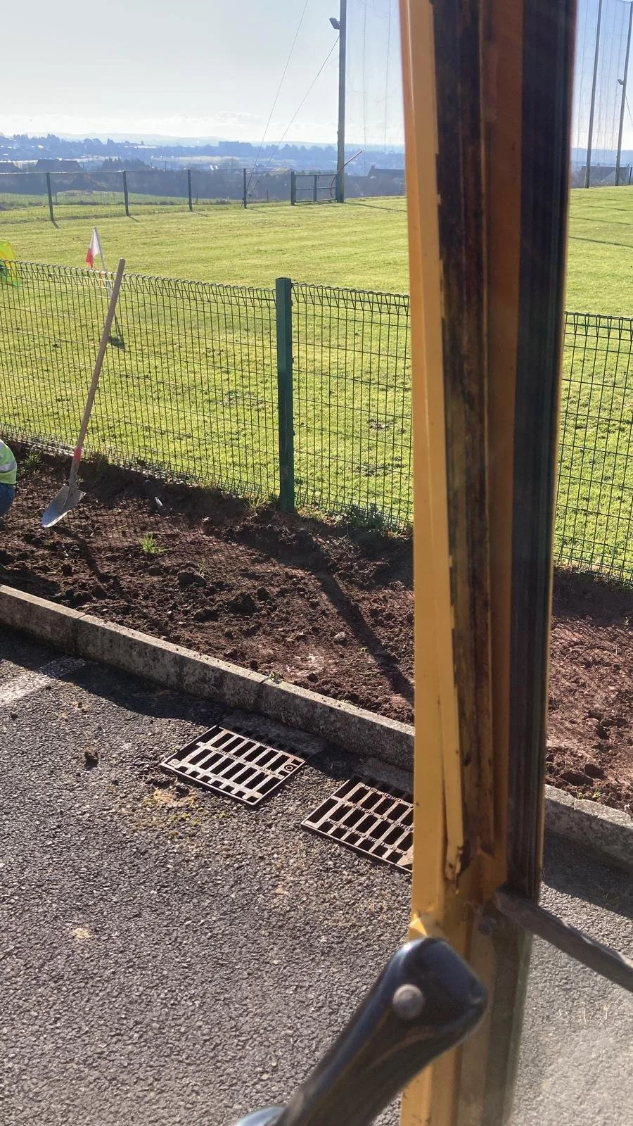 View from inside a bus or tram showing the door frame and a construction worker digging a trench next to a fenced grassy field with a view of distant hills and a city skyline in the background.