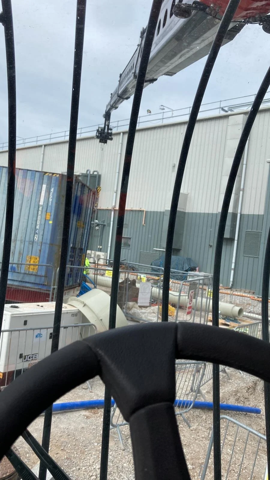 Photo taken from inside a construction vehicle with a steering wheel in the foreground. Behind the vehicle's barred window, there is a construction site with a crane, shipping containers, construction equipment, and workers.