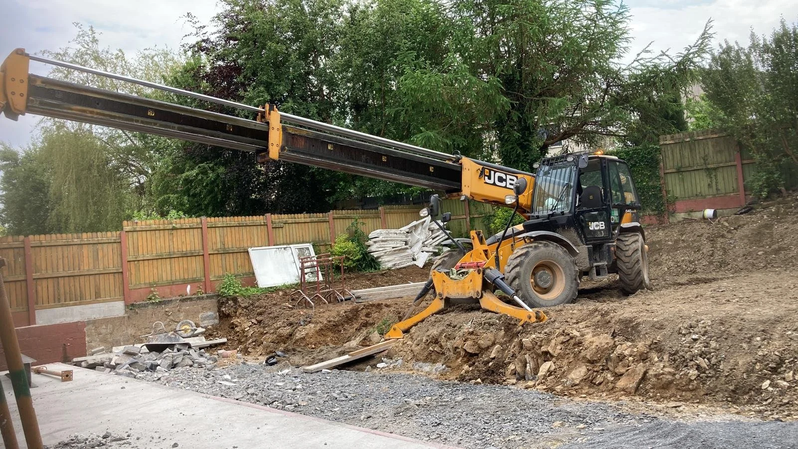 Construction site with excavator moving dirt and rocks, wooden fence and green trees in the background.