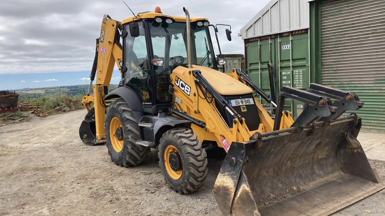 A yellow JCB backhoe loader parked outdoors on a gravel surface with a green storage container and a rolling shutter door in the background.