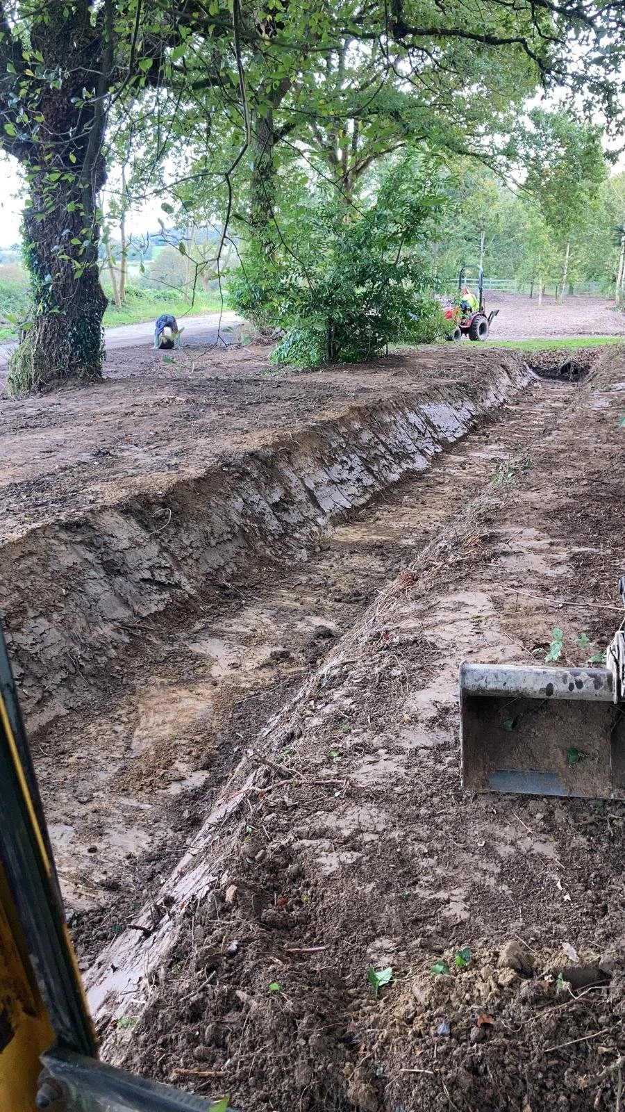 A trench being excavated in a dirt area with trees and greenery around. Construction equipment and workers are visible in the background.