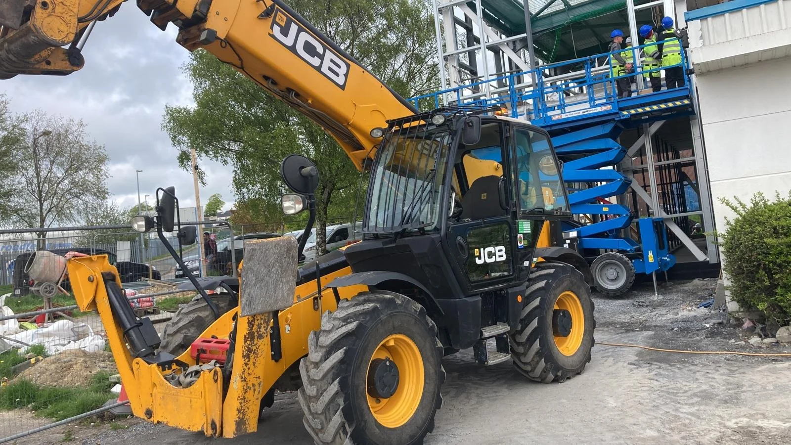 Construction site with a JCB backhoe loader and blue scissor lift, workers in safety gear on the lift platform, building under renovation, overcast sky