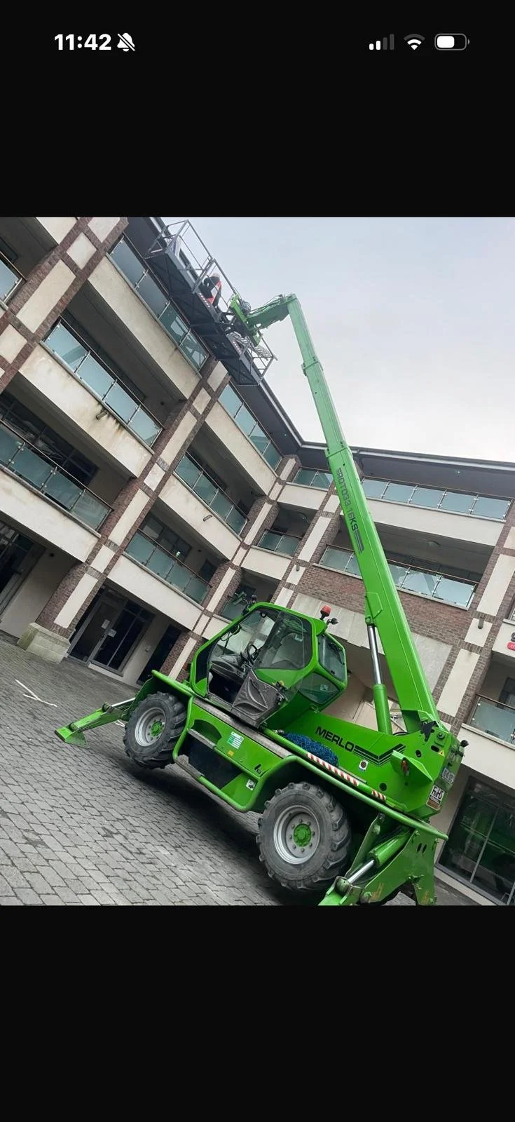 A green telescopic boom lift is extended to the top floor of a multi-story apartment building with brown and beige exterior, glass balconies, and sliding glass doors.
