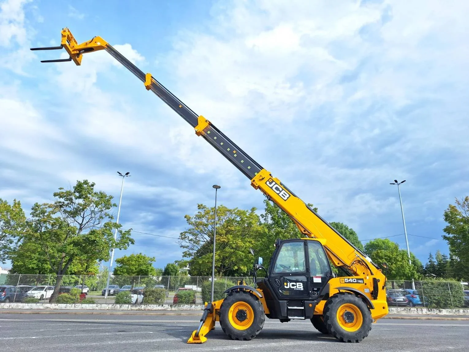 Yellow JCB telescopic boom lift with extended arm, parked on a paved road with trees, a chain-link fence, and parked cars in the background under a partly cloudy sky.