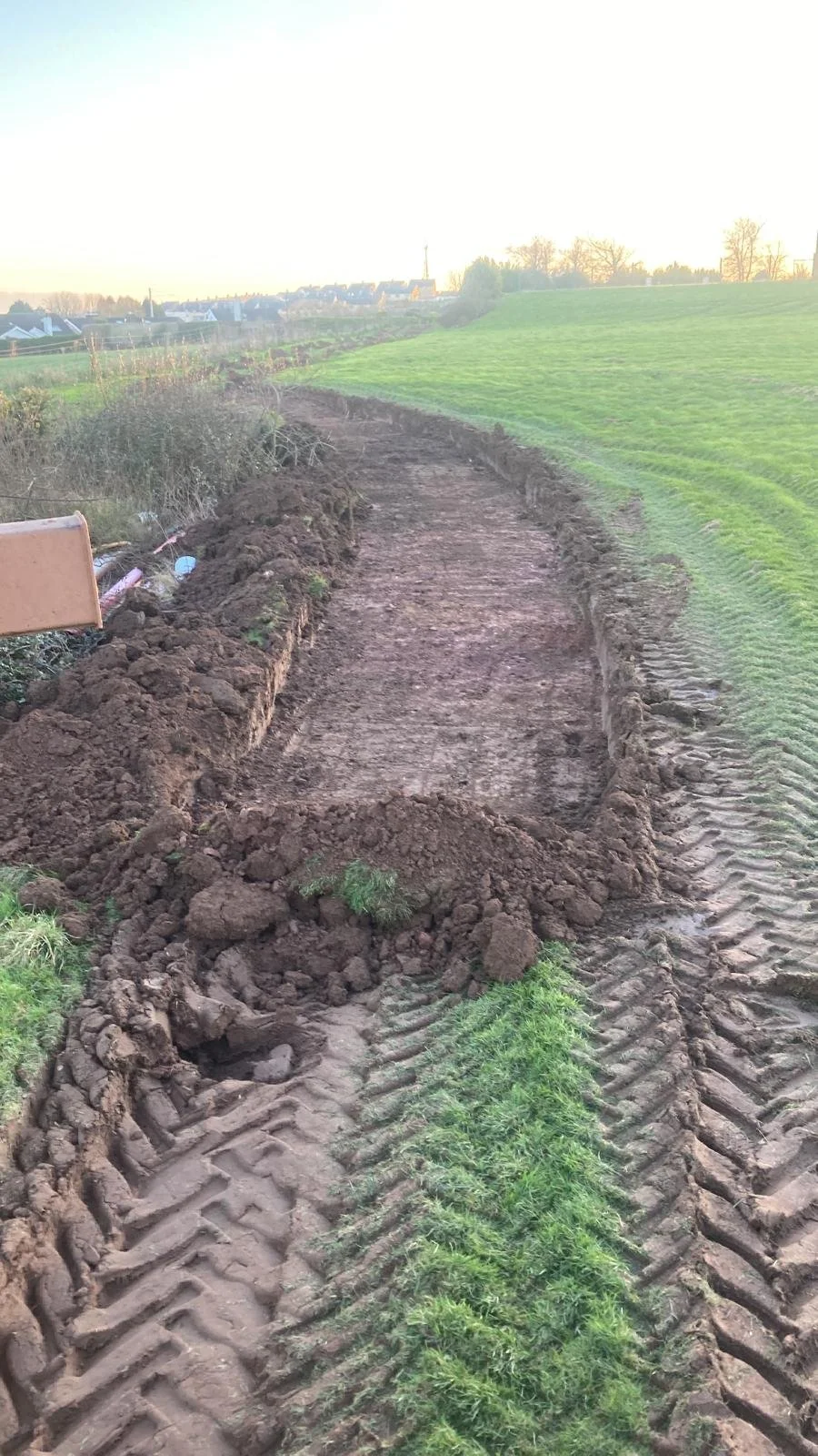 A dirt pathway under construction on a grassy field, with tire tracks from construction machinery visible along the sides, and an open field in the background during sunset.