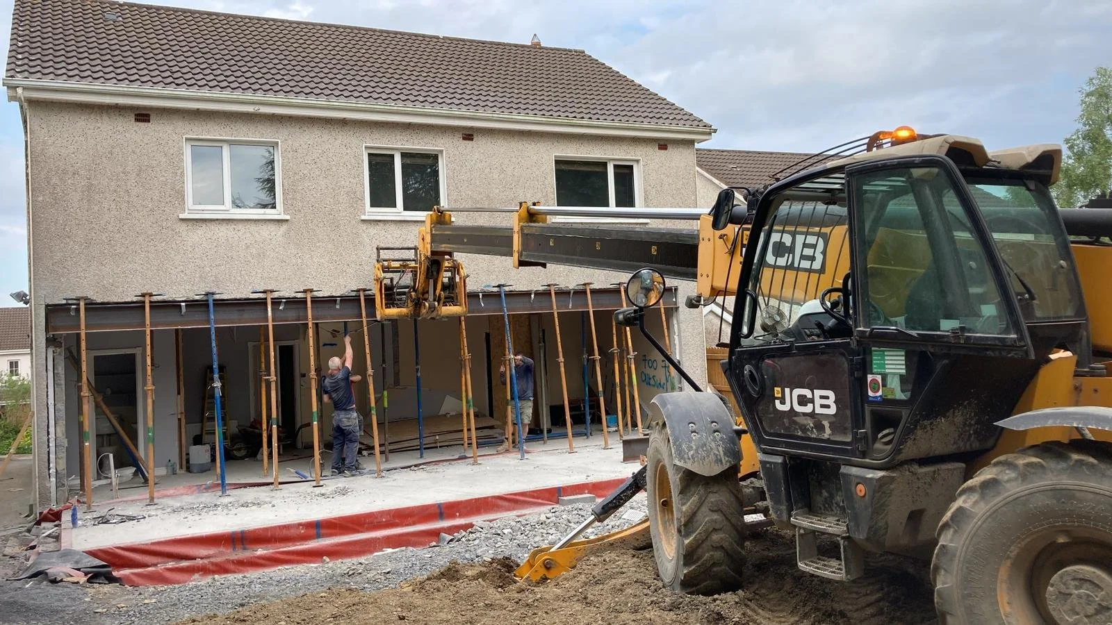 Construction site with workers installing support beams on a building, a large JCB excavator parked nearby, and a residential house in the background.