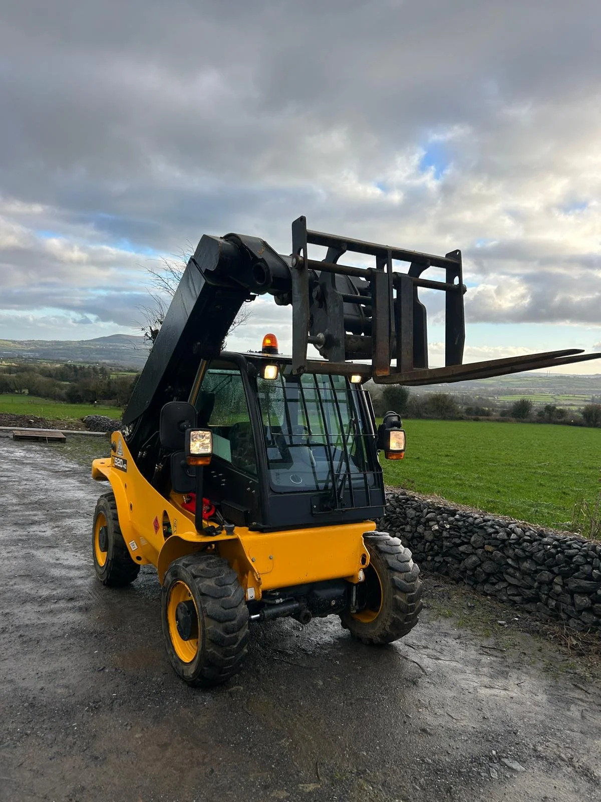 Yellow and black telescopic forklift on wet gravel ground with green fields and cloudy sky in background.