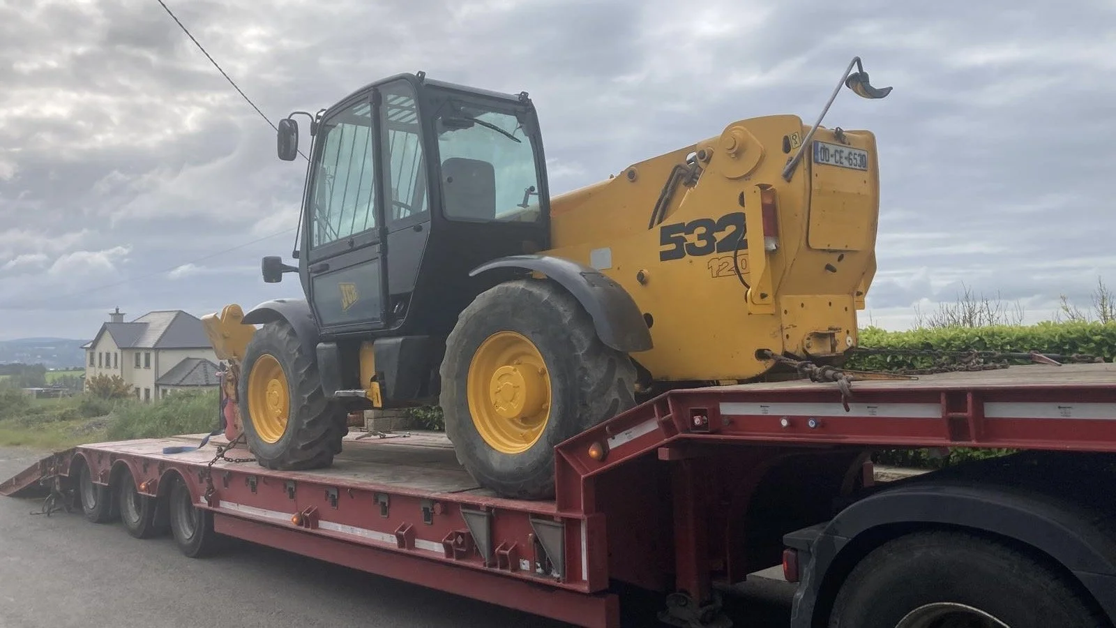 Yellow and black construction vehicle on a red flatbed trailer, parked outdoors with residential buildings and a cloudy sky in the background.