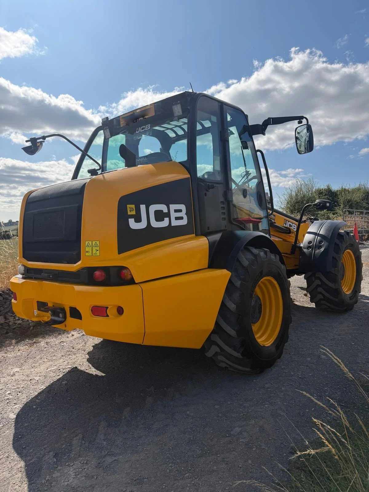 A yellow and black JCB construction vehicle parked on a gravel surface under a partly cloudy sky.