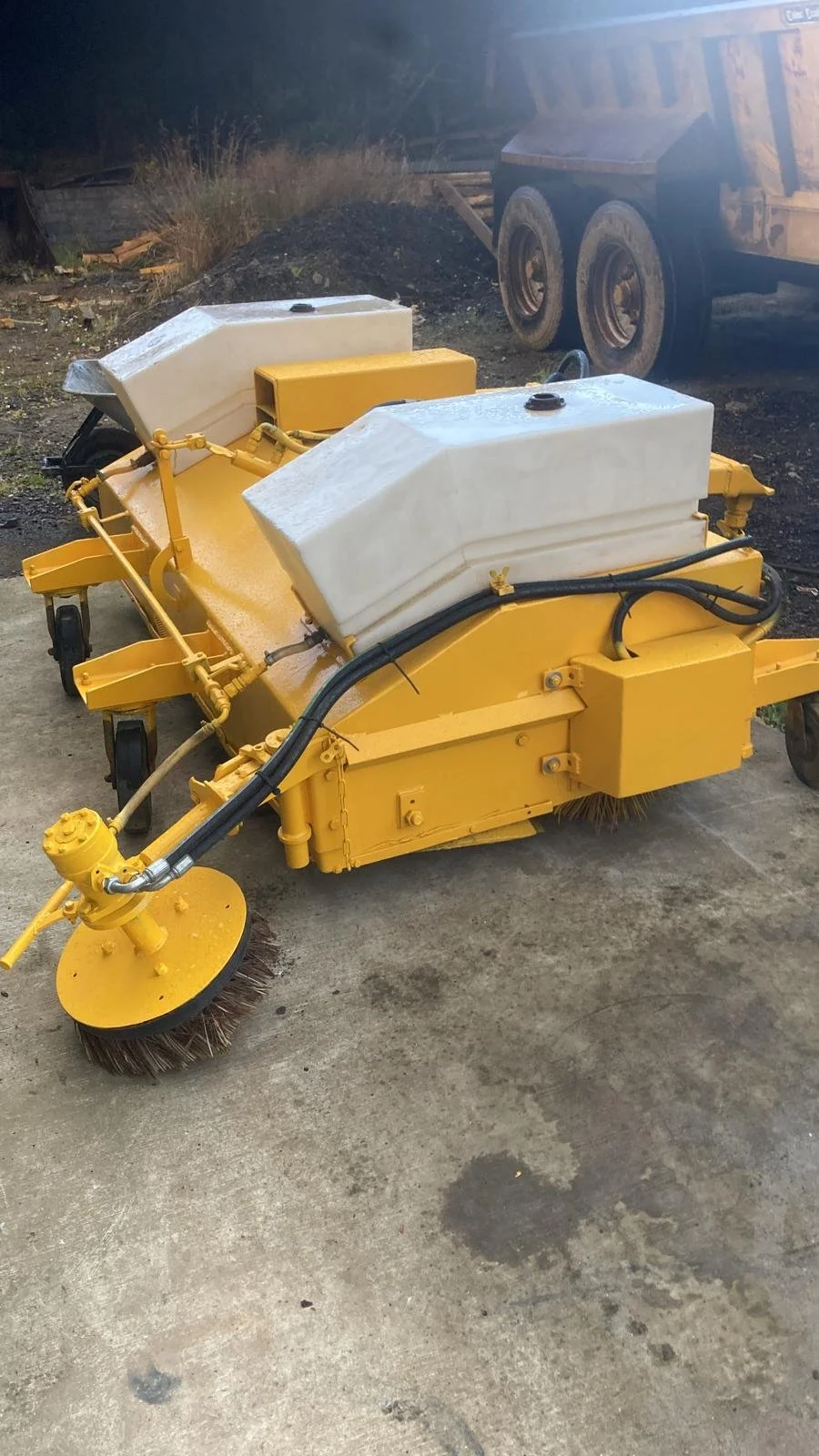 Yellow street cleaning machine with two white water tanks, a side brush, and black hoses, parked on concrete ground with a dirt hill and a large truck in the background.