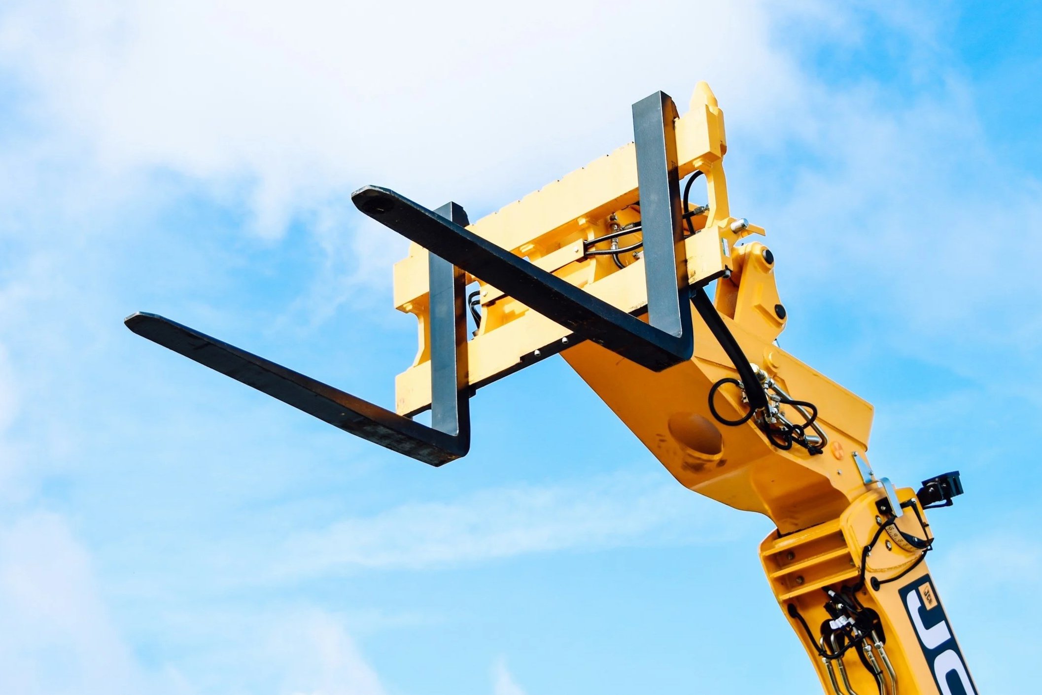 Close-up of a yellow construction crane with black forks against a blue sky with some clouds.