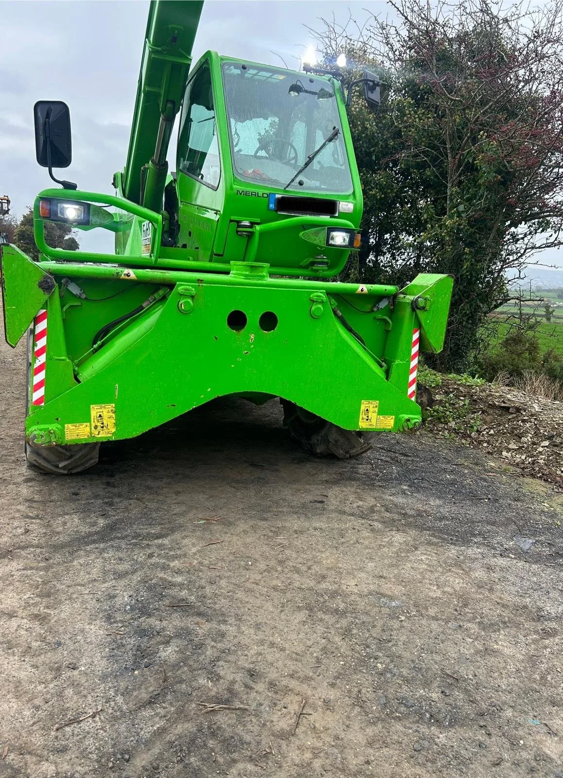 A bright green Merlo aerial lift or cherry picker vehicle on a dirt surface outdoors, with a tree and overcast sky in the background.