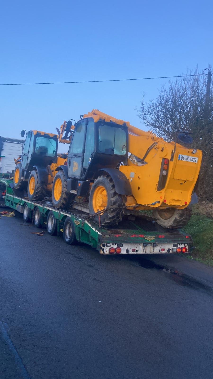 Two yellow construction vehicles on a green flatbed tow truck on a roadside, with clear sky and trees in the background.