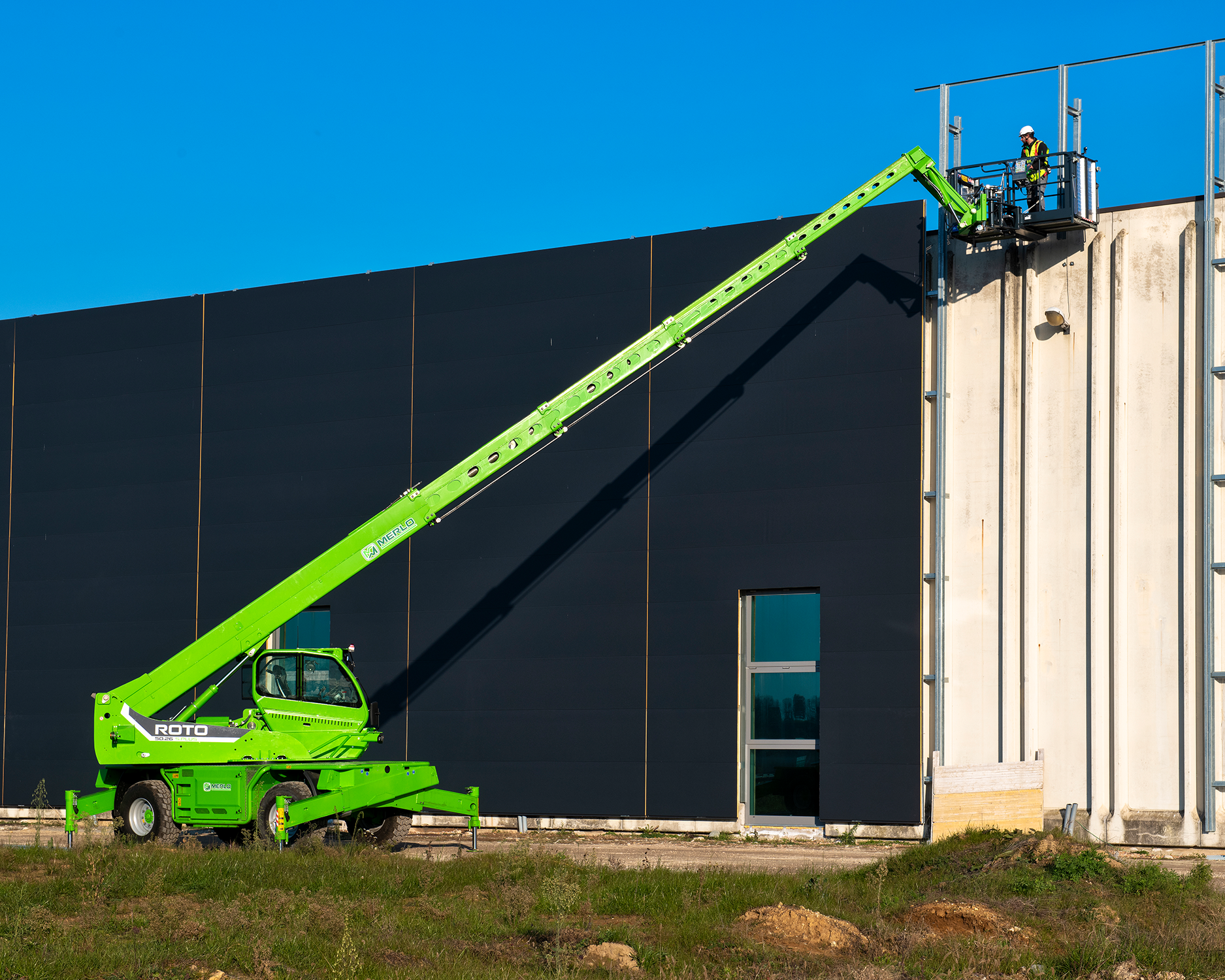 A green Merlo telescopic boom lift with the brand ROTO on its side, extending its arm to assist workers on a high building wall. There are two workers on the platform at the top of the lift, wearing safety gear. The building has dark exterior panels and a small window, set against a clear blue sky.