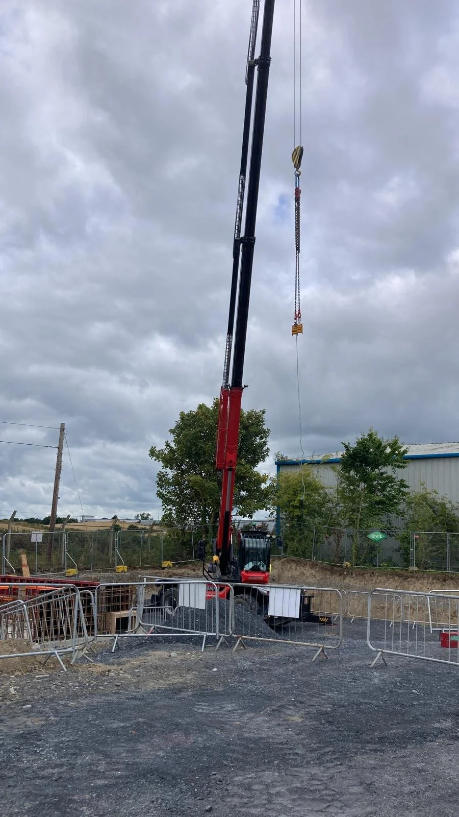 A large construction crane with a black and red body operated at a construction site with gravel ground, surrounded by metal safety barriers, under a cloudy sky.