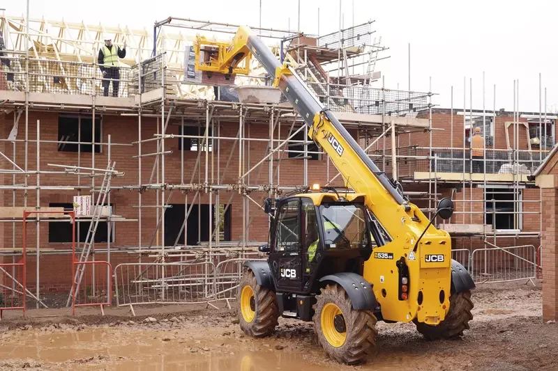 Construction site with a JCB telescopic forklift lifting materials to the upper levels of a building under construction, with workers on-site and scaffolding around the structure.