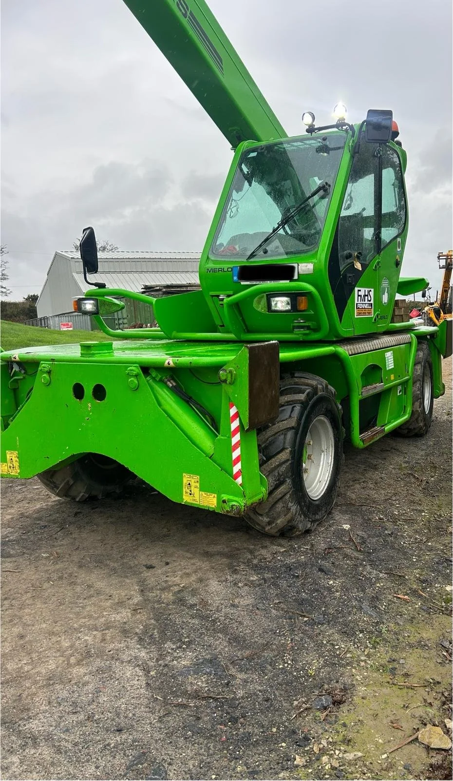 Green telescopic handler or lift truck parked on dirt ground outdoors with a cloudy sky, a building, and some trees in the background.