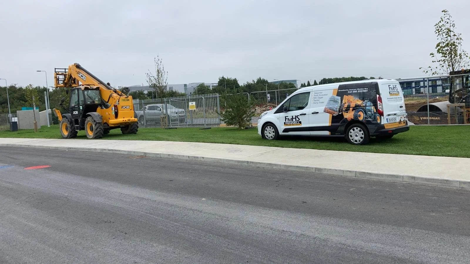 A yellow JCB construction crane parked on a grassy area beside a sidewalk, with a white FmHS Fennell service van nearby, and a construction site with fencing and equipment in the background.