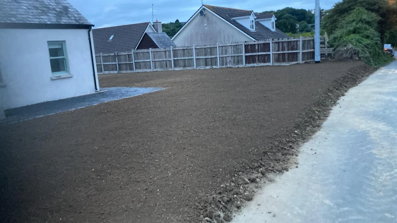 A residential backyard with a dirt lawn, a white house on the left, a wooden fence, and neighboring houses in the background. The scene appears to be taken during dusk or early evening.