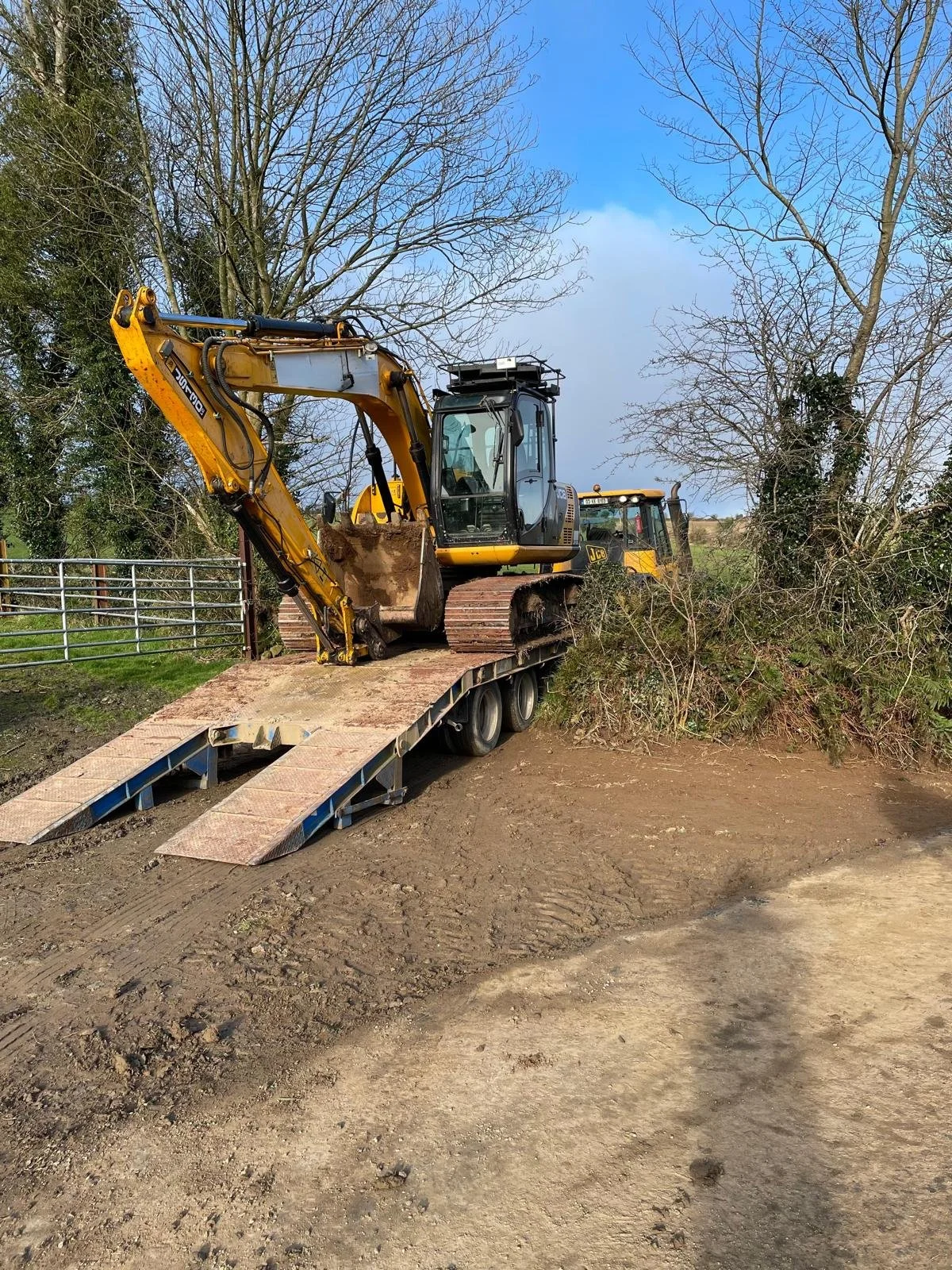 A yellow excavator on a trailer being prepared for transport or unloading, with trees and a partly cloudy sky in the background.