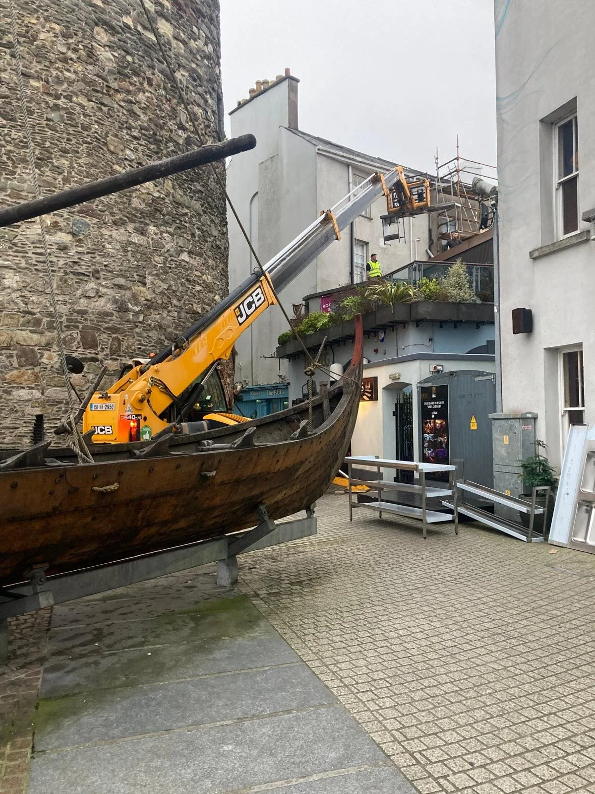 A boat with a yellow crane on it, lifting workers in a bucket to the top of a building under construction or renovation. The scene is set in an alley with old stone walls and modern surrounding buildings.