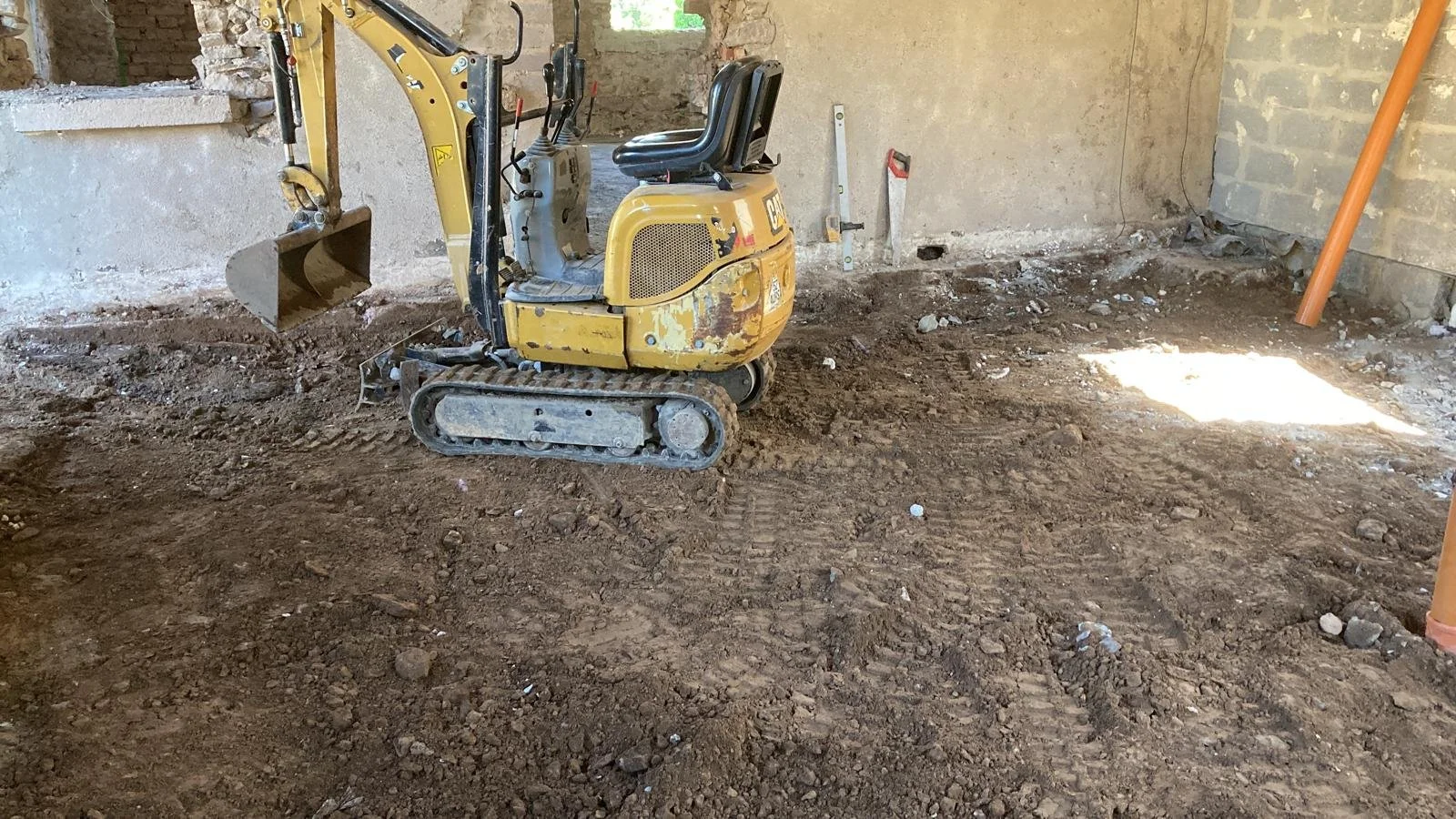 A small, yellow Caterpillar excavator on a dirt floor inside an unfinished room with brick and concrete walls. There are construction tools and equipment around.