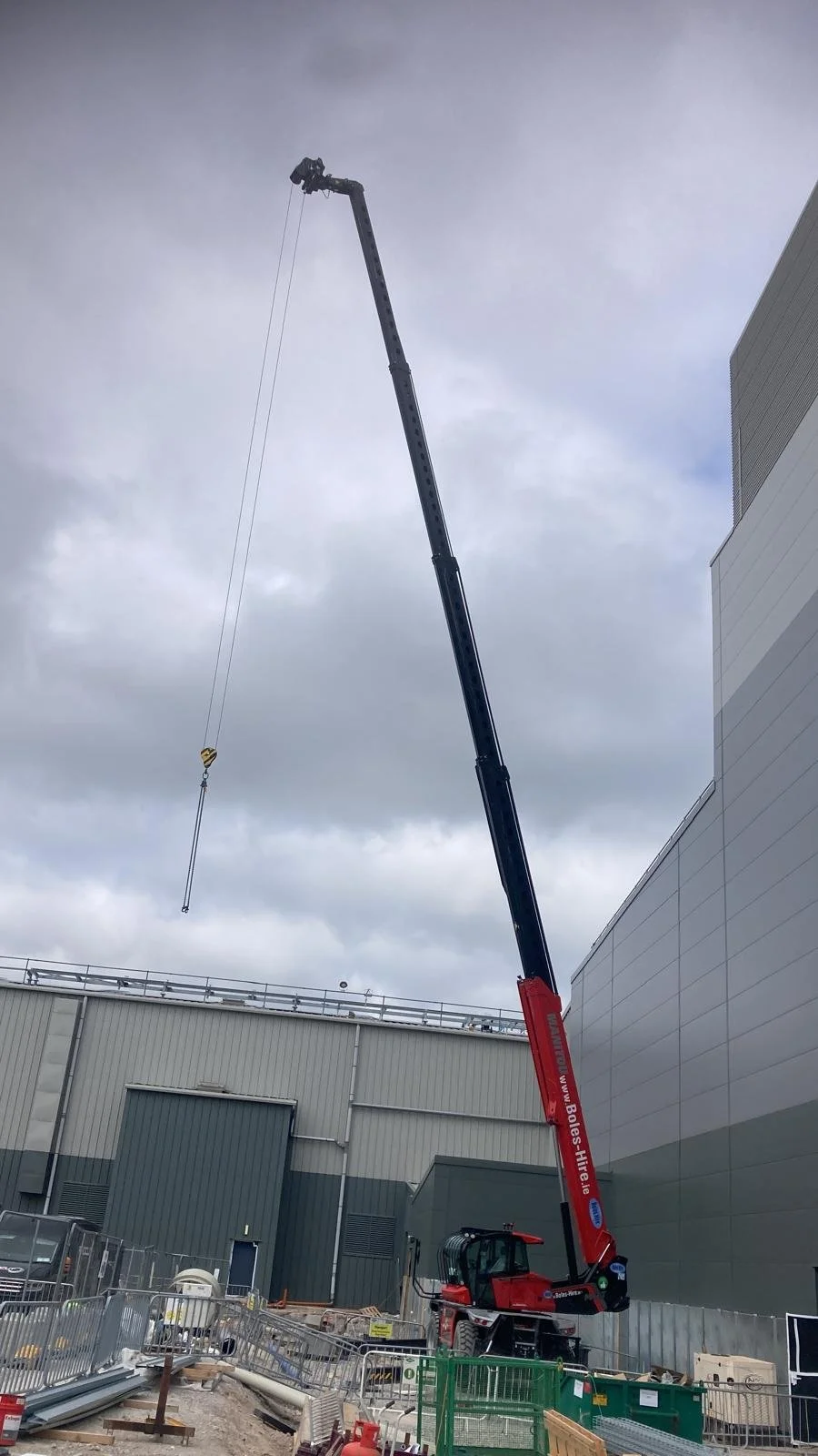 Construction crane lifting a long pole at a construction site with large industrial buildings and cloudy sky in the background.