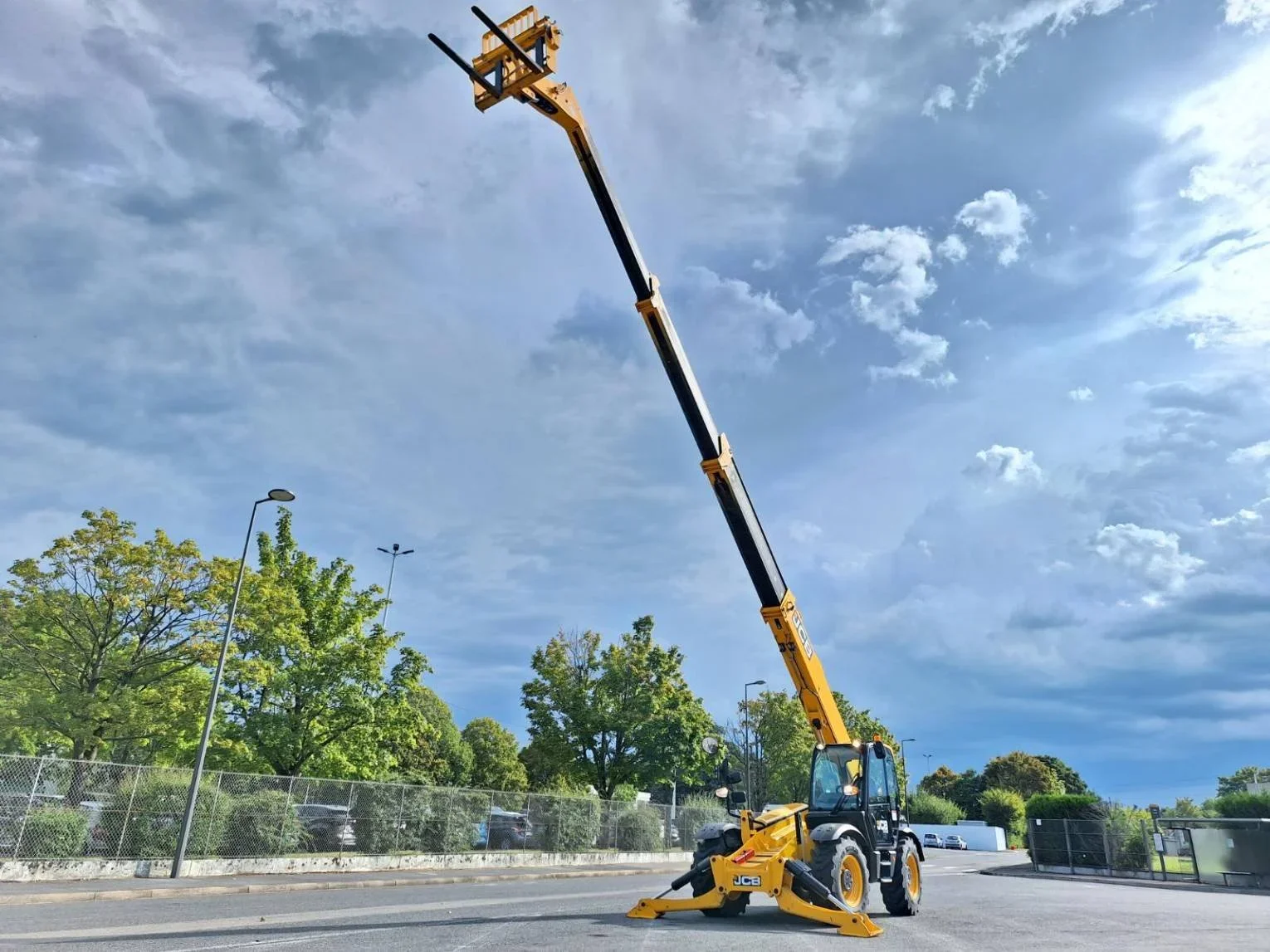 A yellow telescopic boom lift extends upward over a paved area with green trees, streetlights, and a blue sky with clouds in the background.