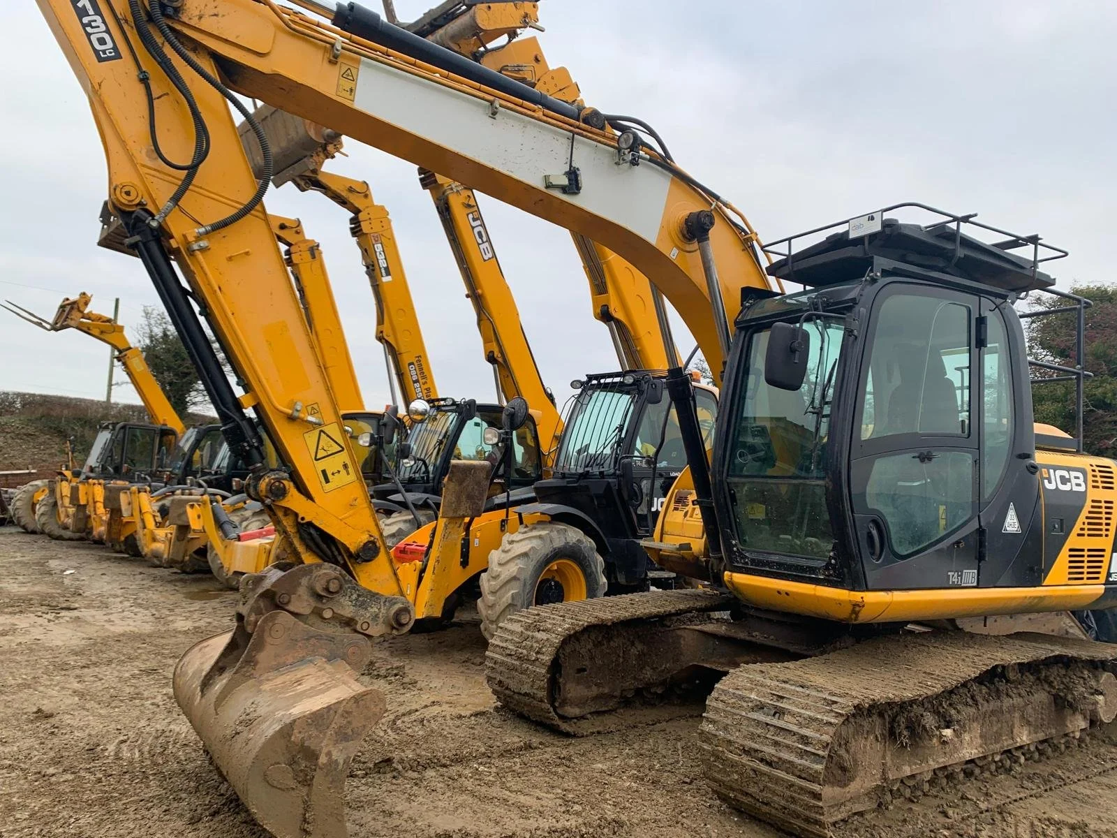 A row of yellow and black JCB excavators lined up on a construction site with dirt ground, under a cloudy sky.