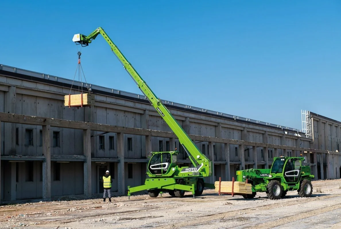 Construction site with a green telescopic crane lifting wood pallets, a worker in a yellow safety vest and helmet, and a forklift nearby, all set against a concrete building and clear blue sky.