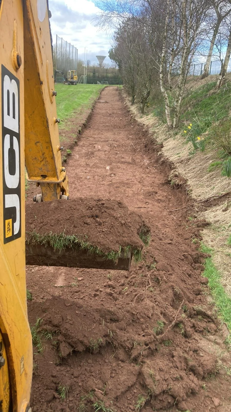 A yellow excavator clearing a dirt pathway beside a grassy area with trees, with a sports field and a silo in the background under a cloudy sky.