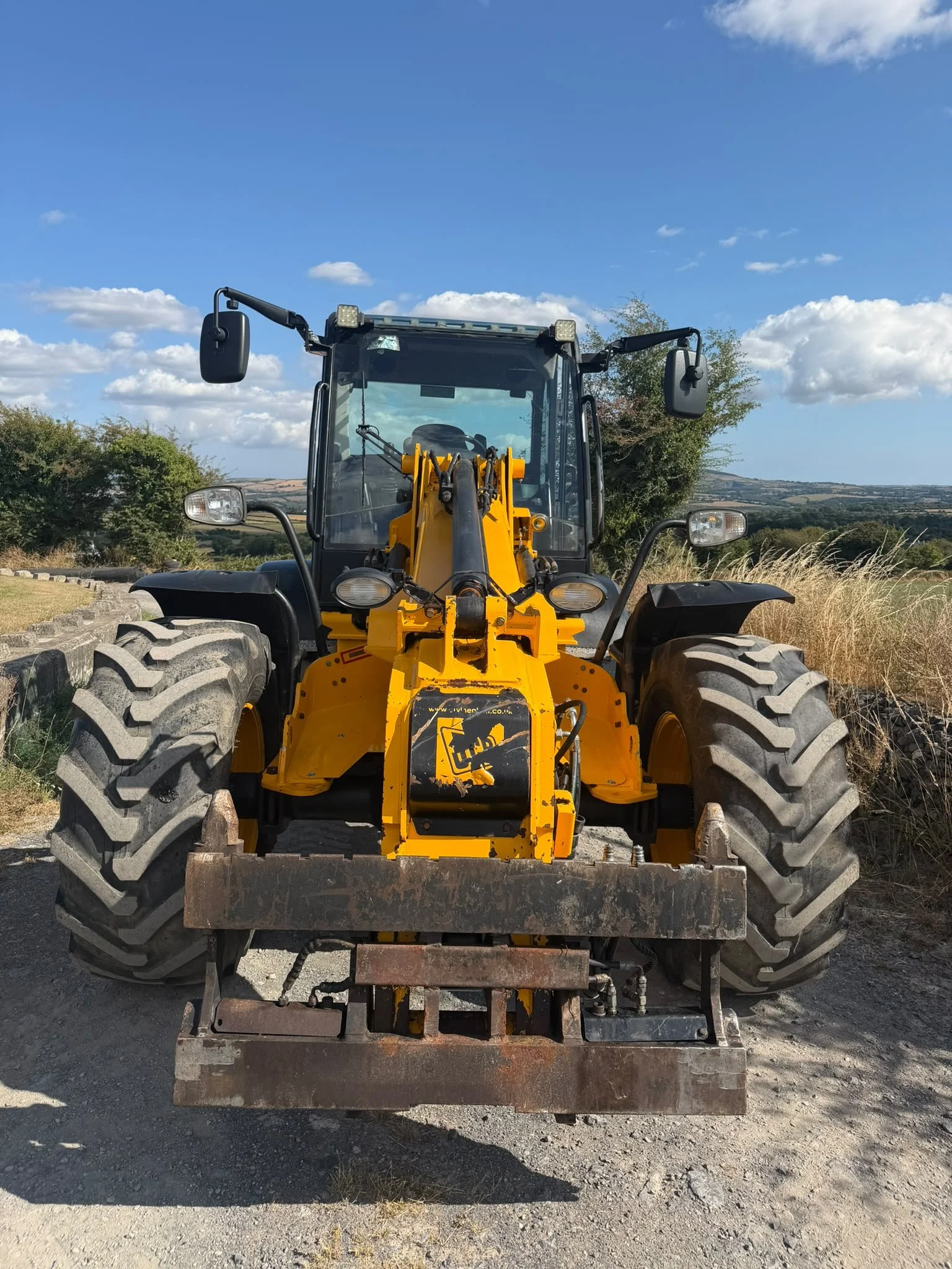 Front view of a yellow construction loader with large tires, parked outdoors under a blue sky with scattered clouds, with trees and rolling hills in the background.
