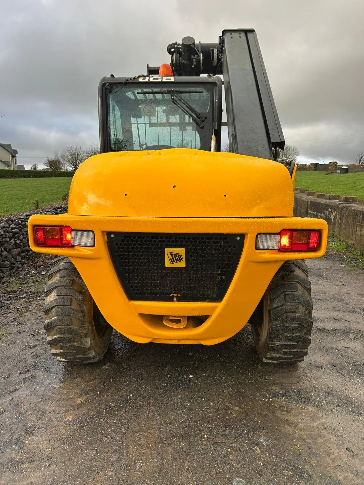 Front view of a yellow JCB construction loader with large tires and a black lifting arm, parked on a gravel surface with a grassy hill and cloudy sky in the background.