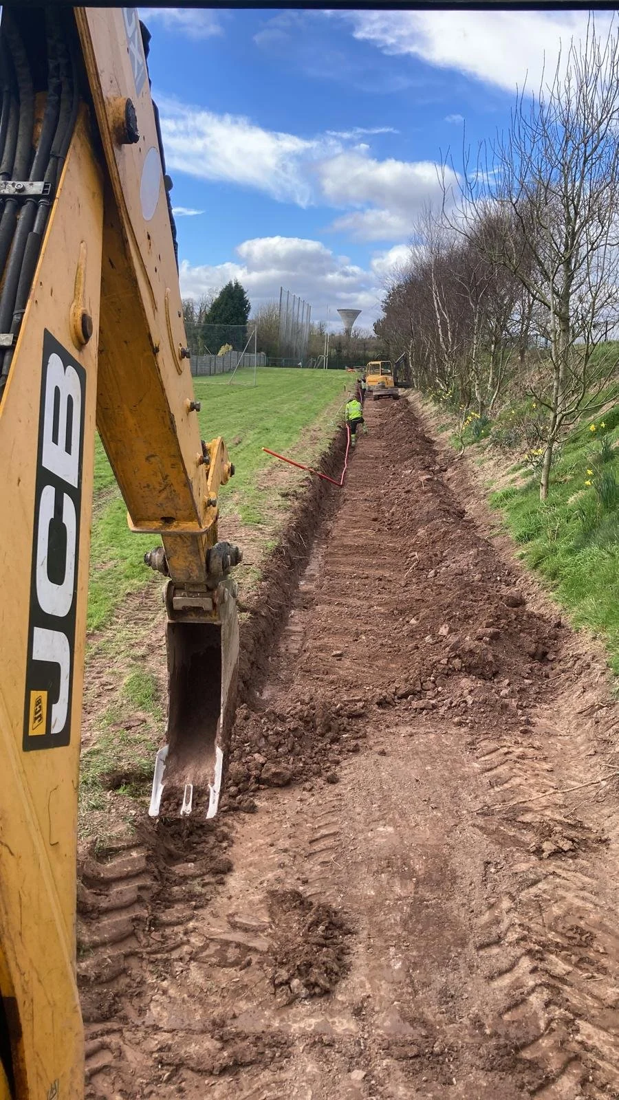 Construction workers and a JCB excavator digging a trench along a grassy area next to trees under a partly cloudy sky.