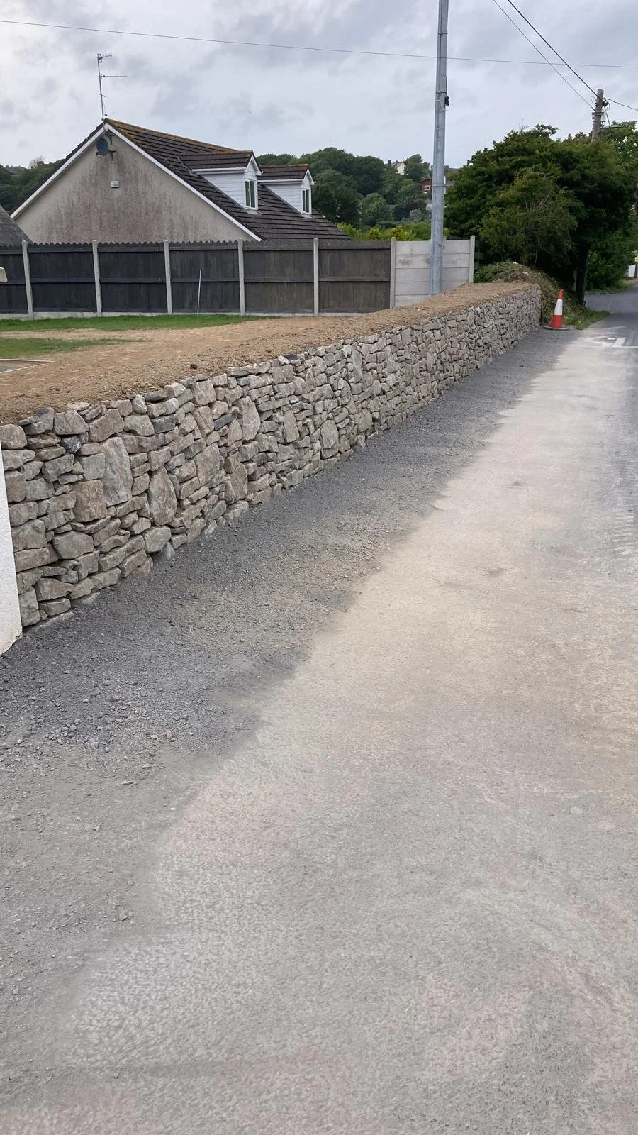A stone wall runs along the edge of a dirt and gravel road in a residential area. Behind the wall, there is a house with a sloped roof and dormer windows, and a wooden fence. The sky is cloudy.