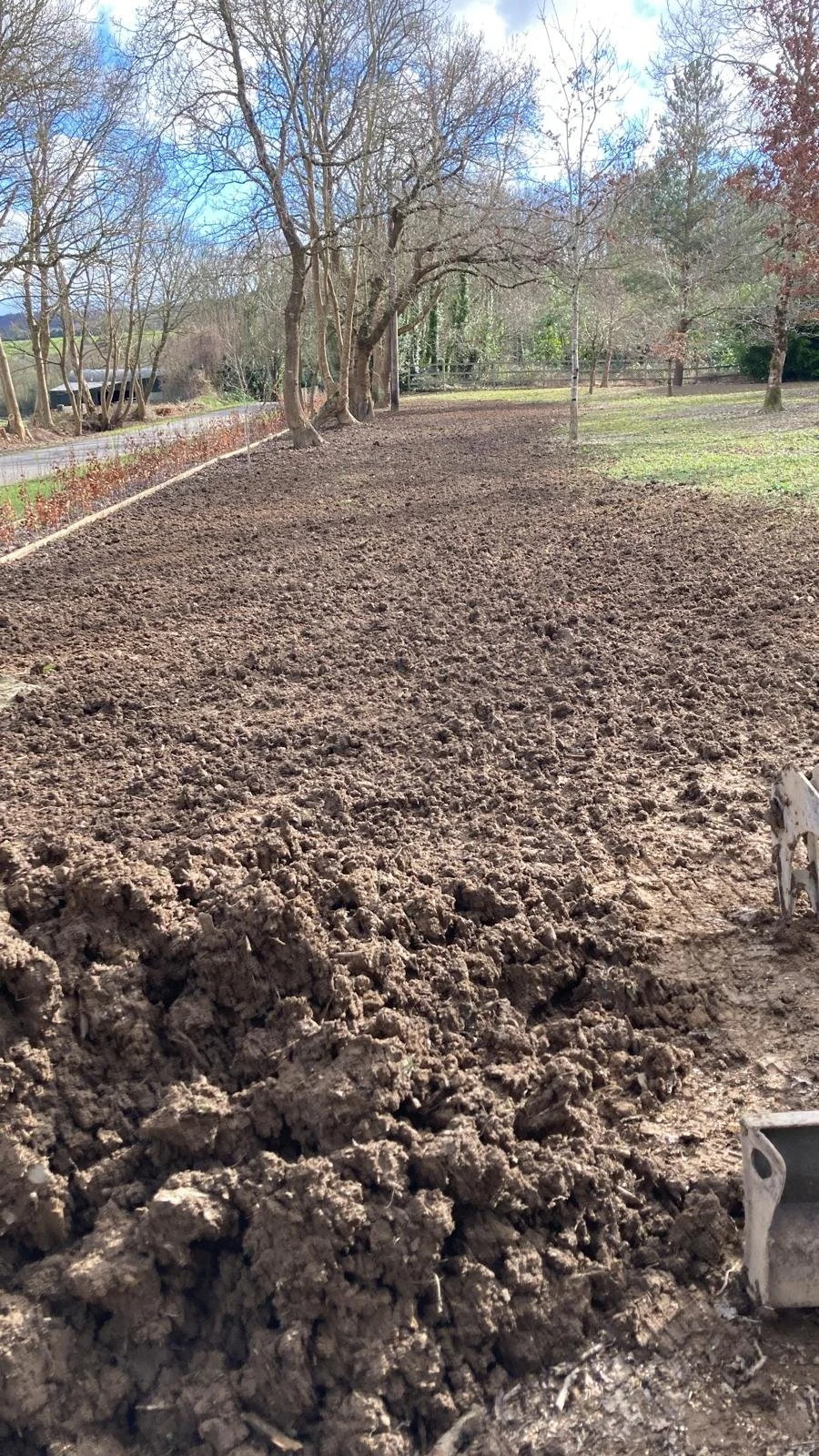 A dirt garden bed being prepared for planting with trees lining the background and a blue sky with some clouds.