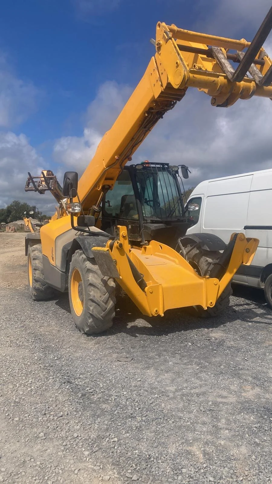 Yellow telescopic forklift with extended arm parked on gravel, with a white van nearby, under partly cloudy sky.