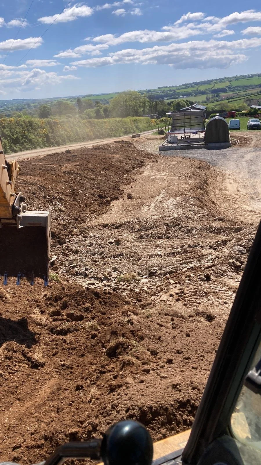 View from construction equipment showing a dirt road being prepared with packed earth, with rolling green hills and a partly cloudy sky in the background.