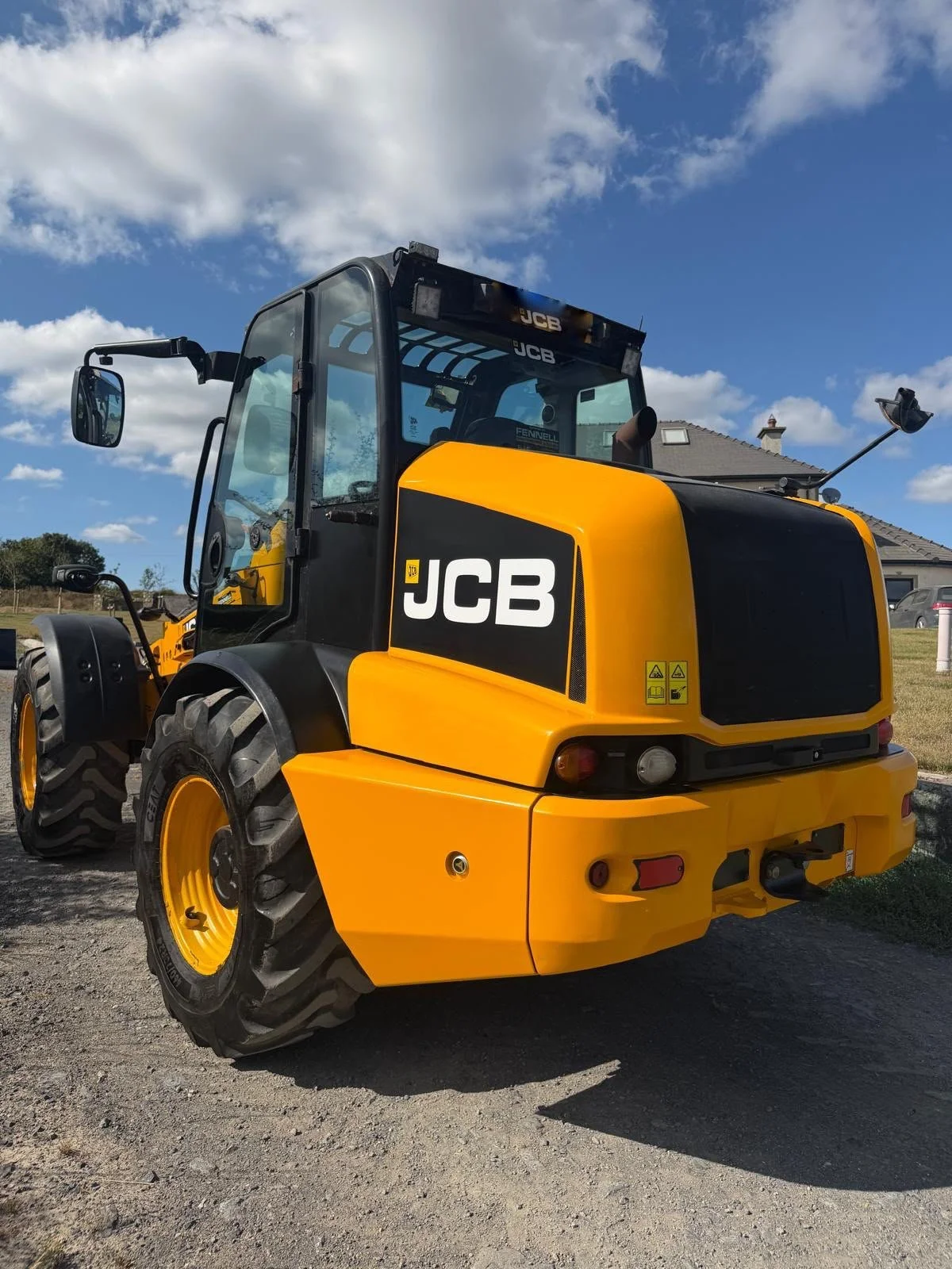 A yellow and black JCB compact wheel loader parked outdoors on a dirt surface, with a house and a blue sky with clouds in the background.