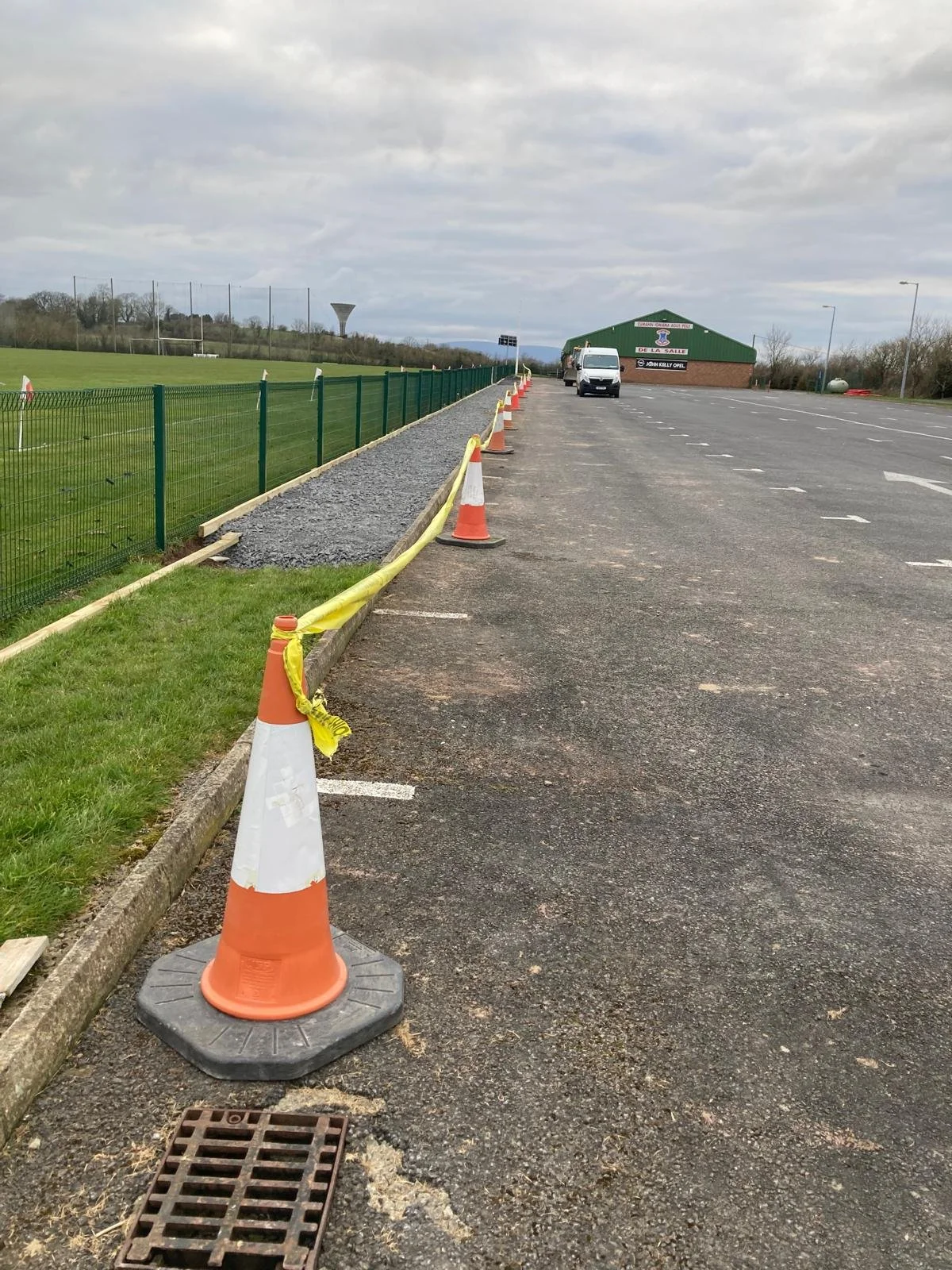 A parking lot with orange and white traffic cones and yellow caution tape along the curb. There's a patch of gravel being prepared on the side, next to a green fence, with a sports field behind it. A white van is parked near a green building in the d