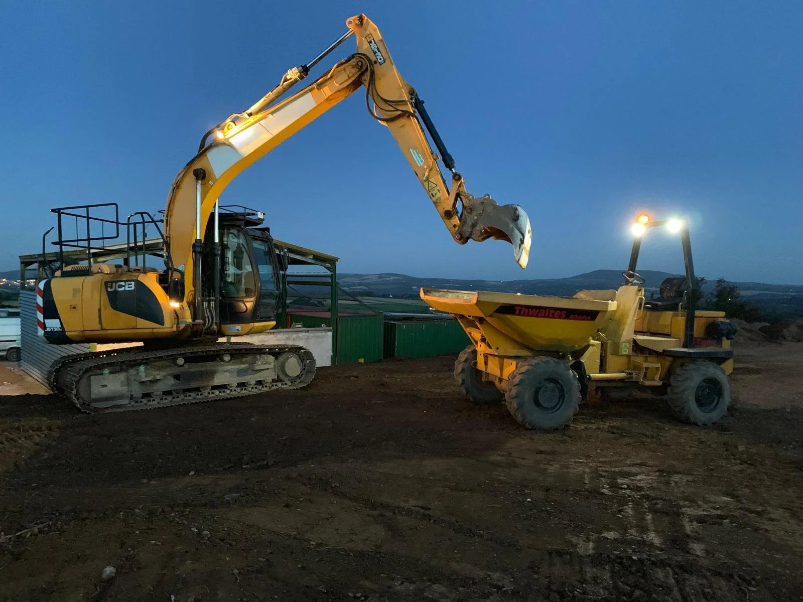 Construction site with a blue sky at dusk showing a yellow excavator transferring soil into a yellow dump trailer.