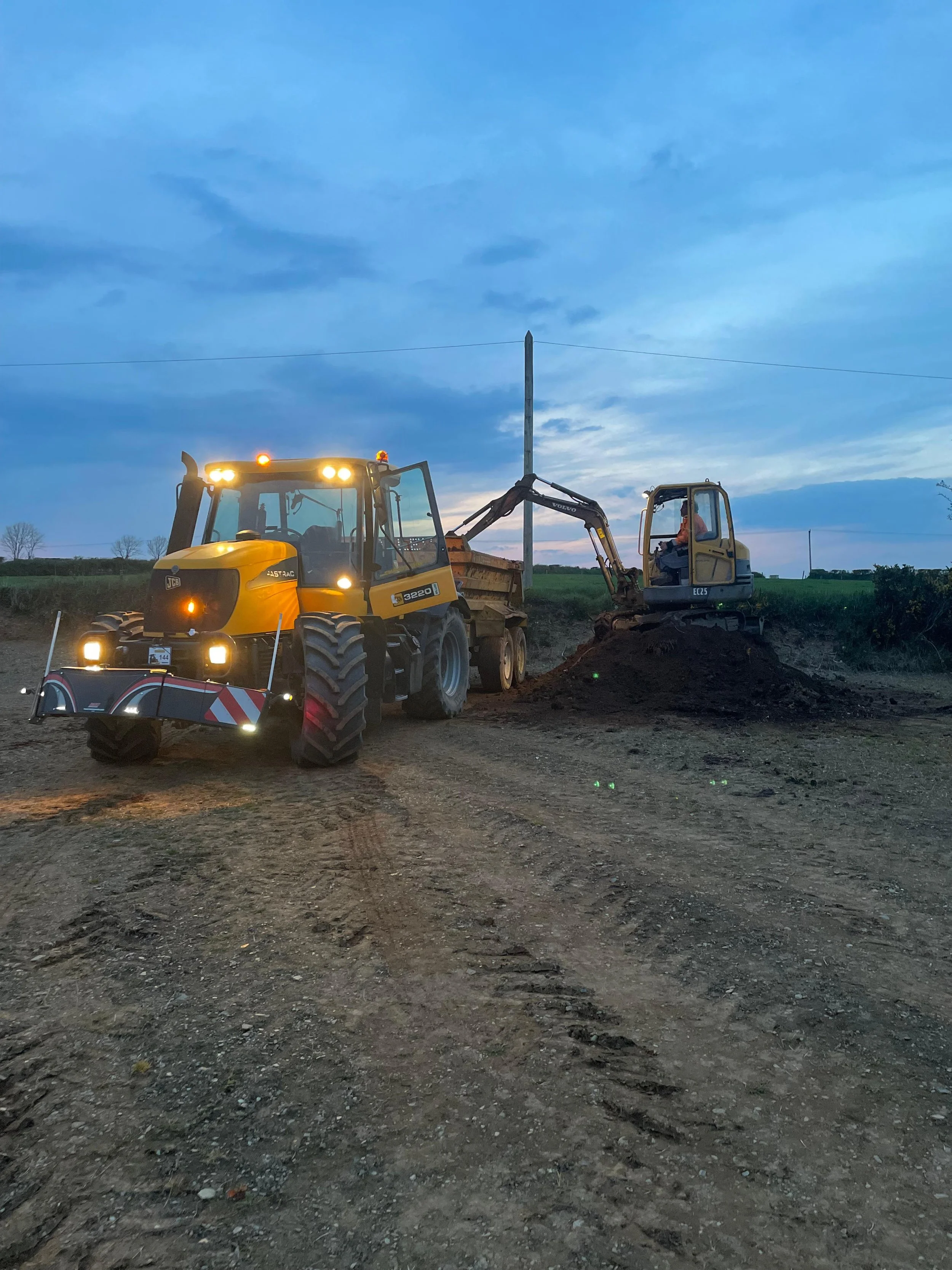 Construction site showing two yellow construction vehicles working on the soil during dusk, with a telephone pole and a partly cloudy sky in the background.