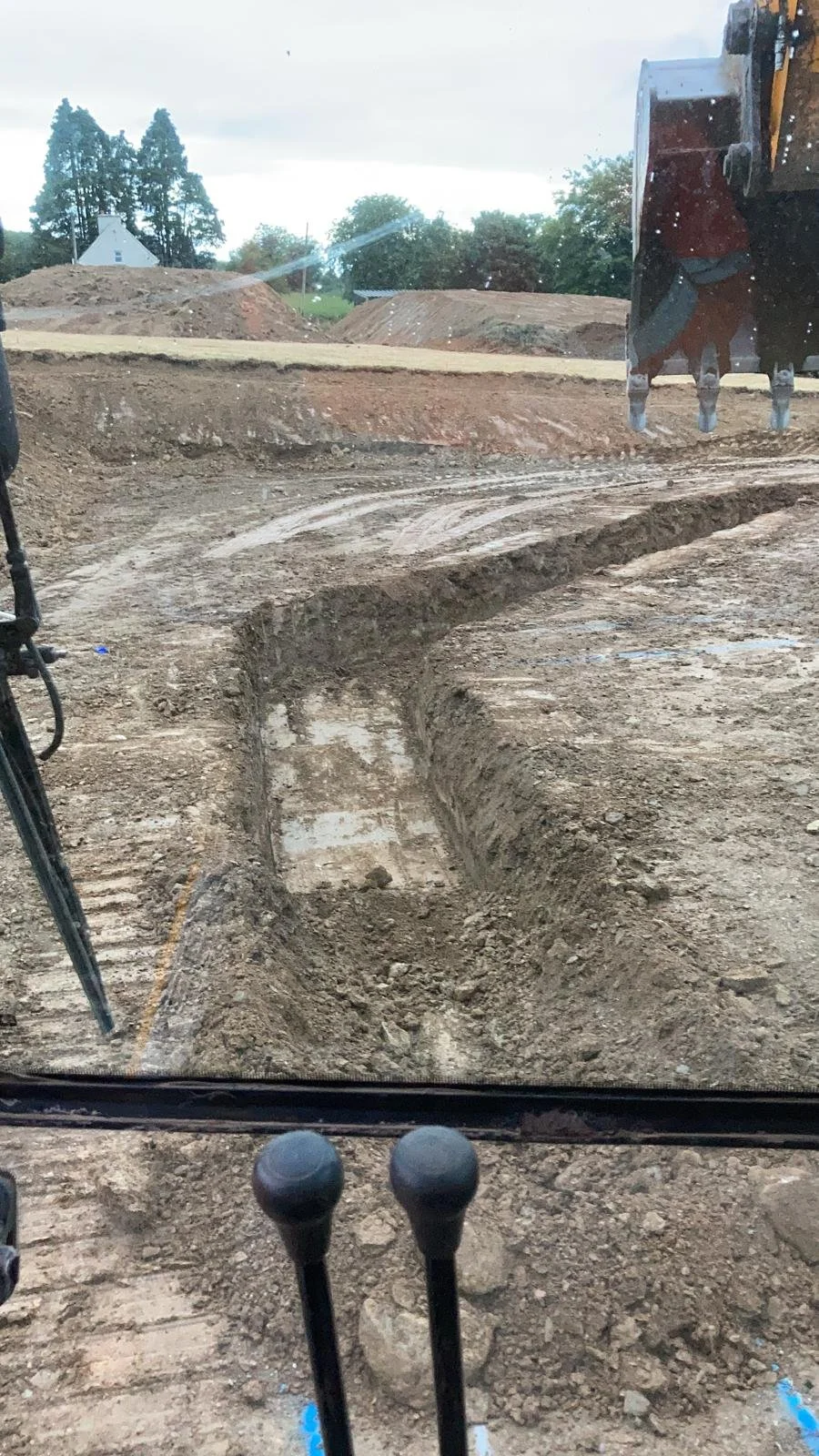 Construction site with excavated trenches, dirt mounds, and construction equipment visible through the window of a heavy machinery cab during daytime.