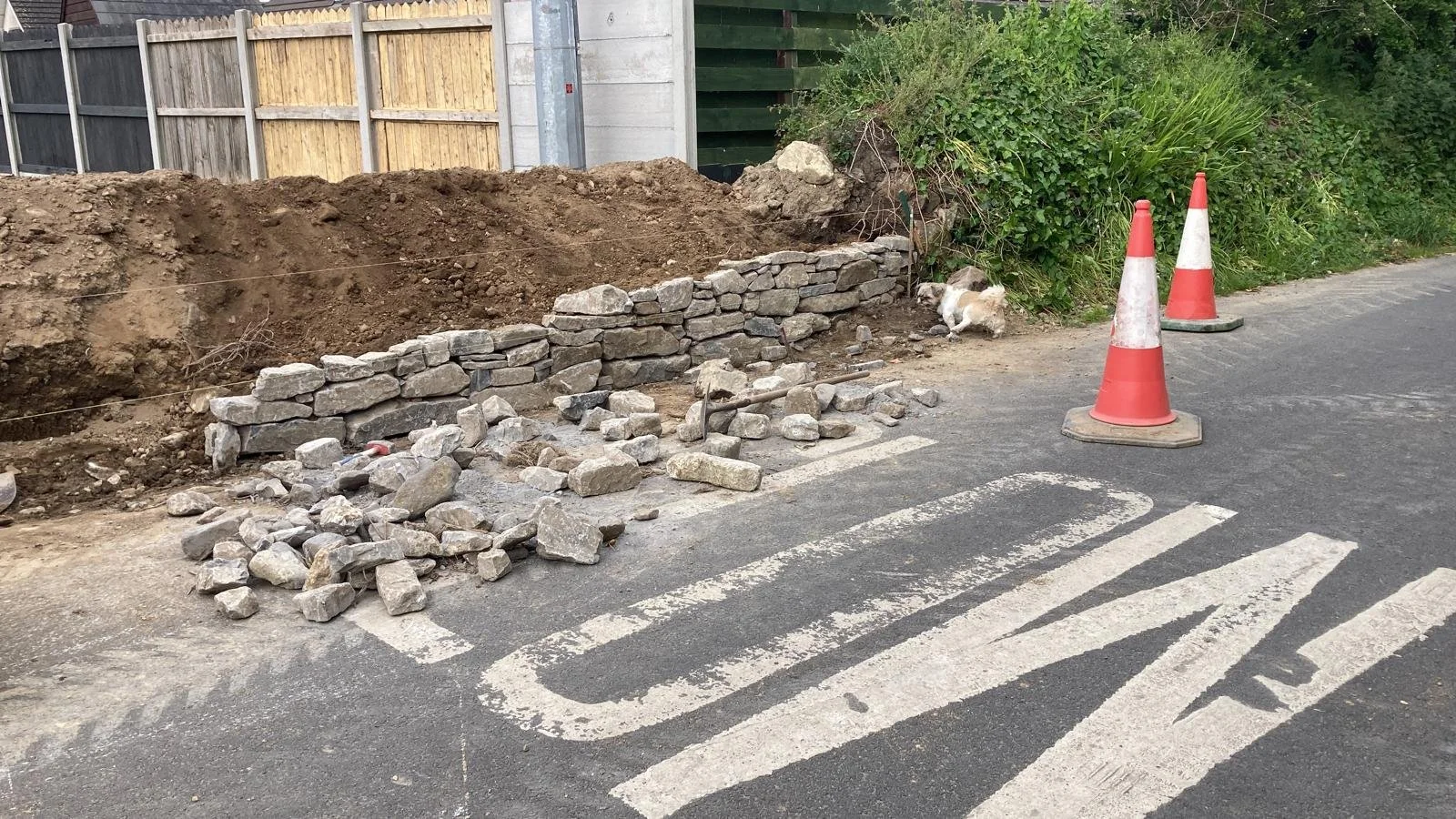 Construction site with a stone wall being built, piles of rocks, and orange traffic cones on a street with a tire tread marking.