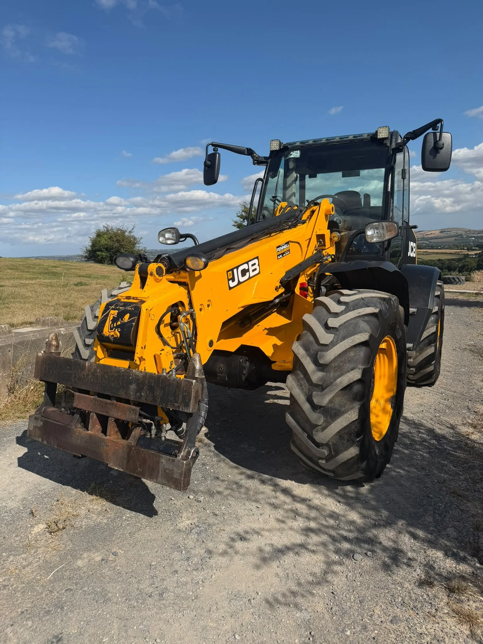 A yellow JCB construction loader parked on a dirt area in a rural landscape with trees, grass, and hills under a blue sky with some clouds.